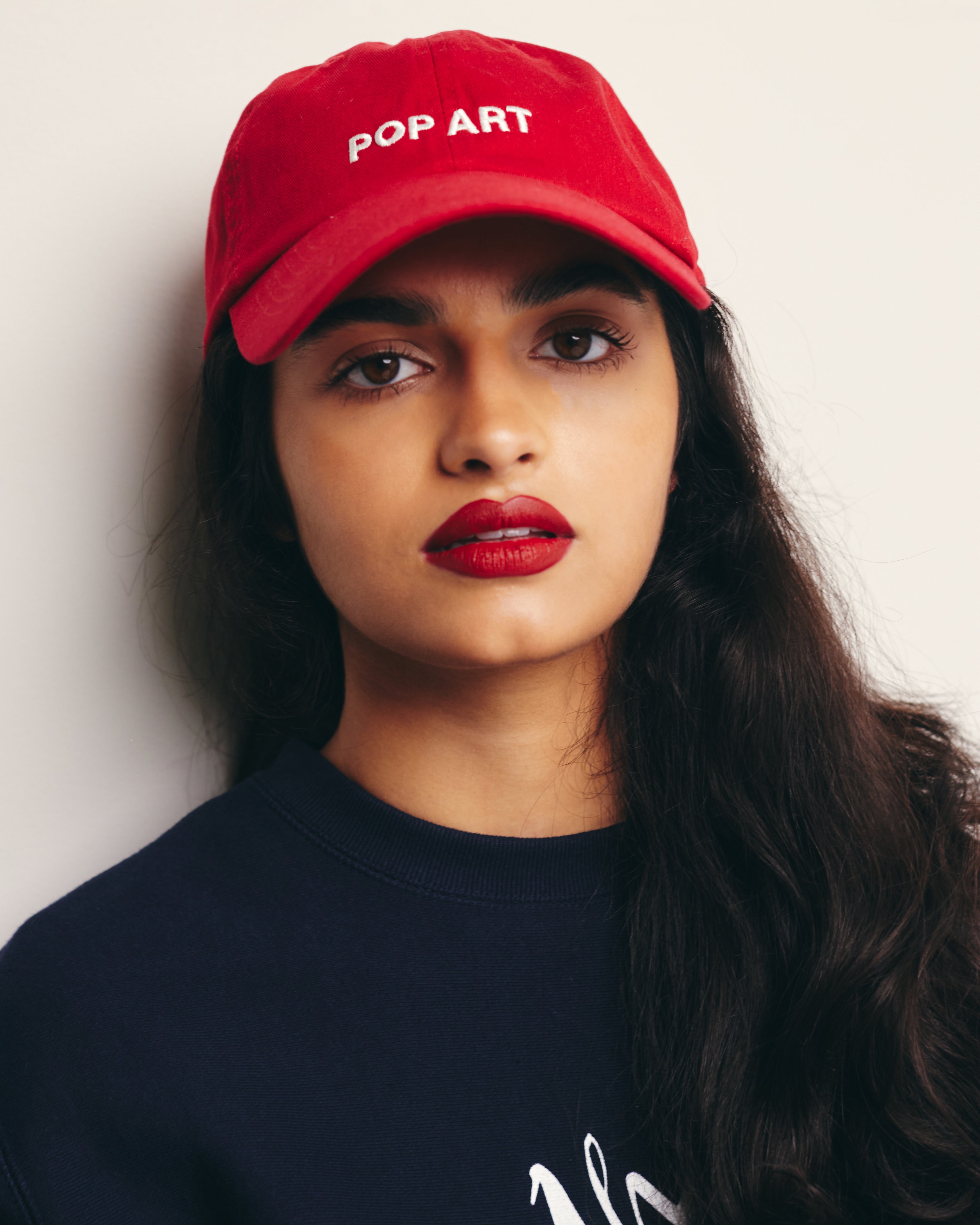 Mira Bhat wears Red Baseball Cap / Dad Hat with Pop Art Movement Text Embroidery on Front. 100% Cotton.